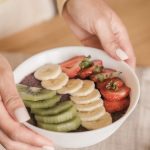 Delicious smoothie bowl topped with fresh fruits like kiwi, banana, and strawberries, held by manicured hands.